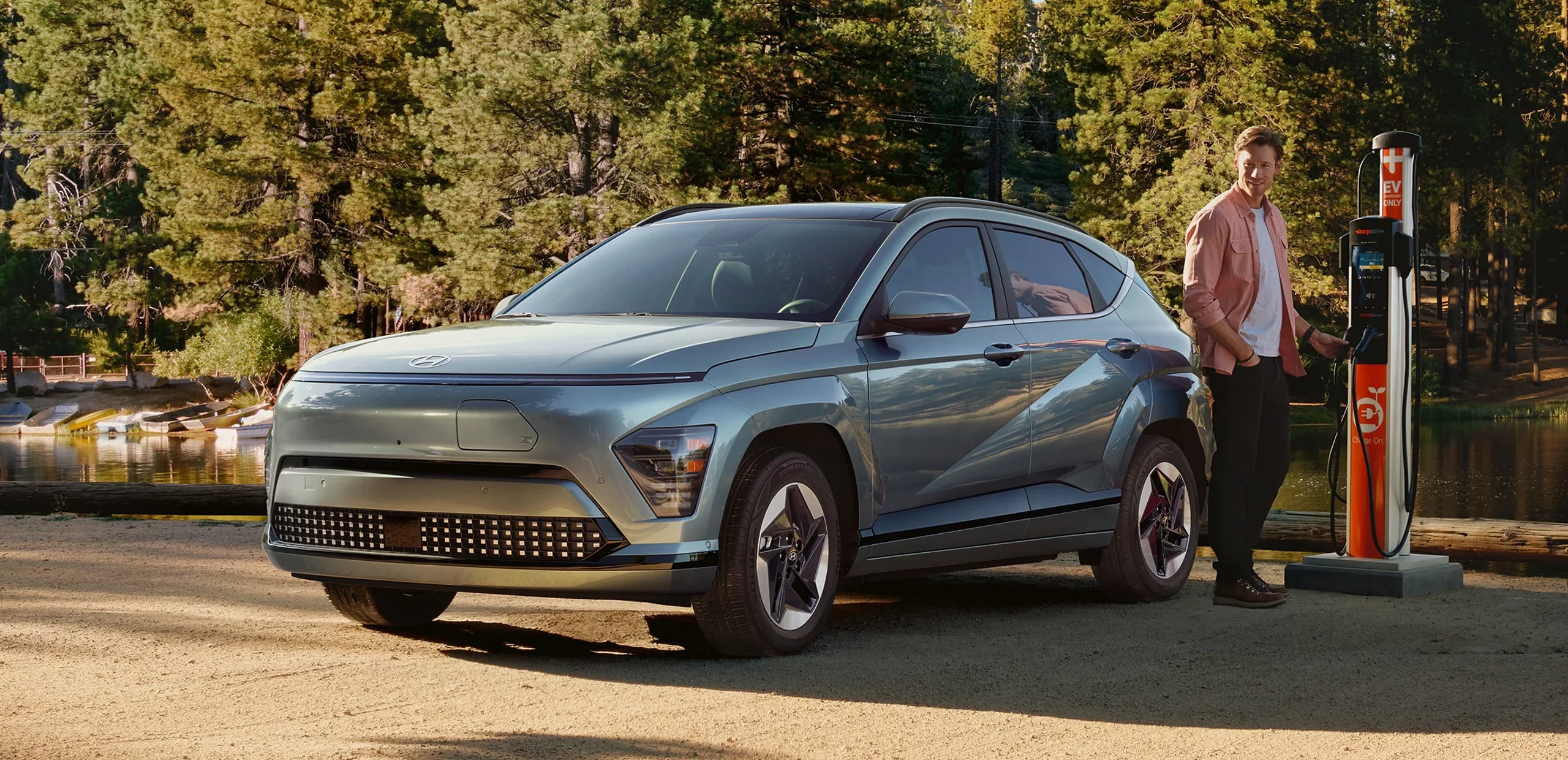 A man plugs a charger into a light blue Kona Electric SUV parked on a dirt road beside a lake surrounded by trees.